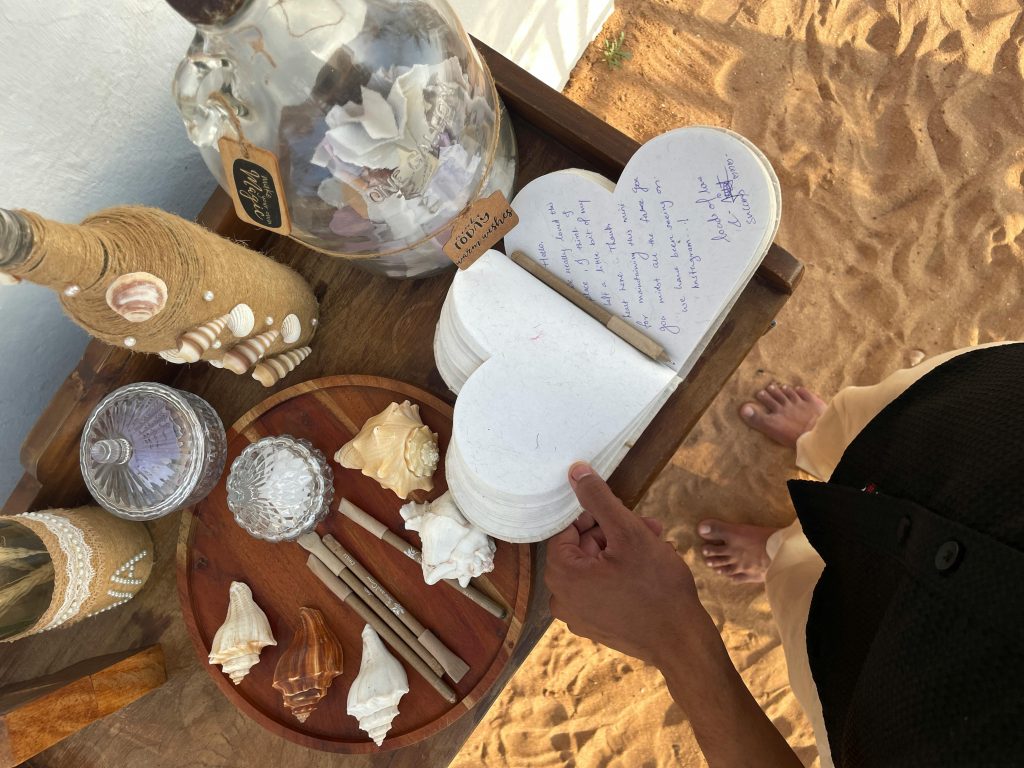 A beachside table with seashell decor and a heart-shaped guestbook on sand.